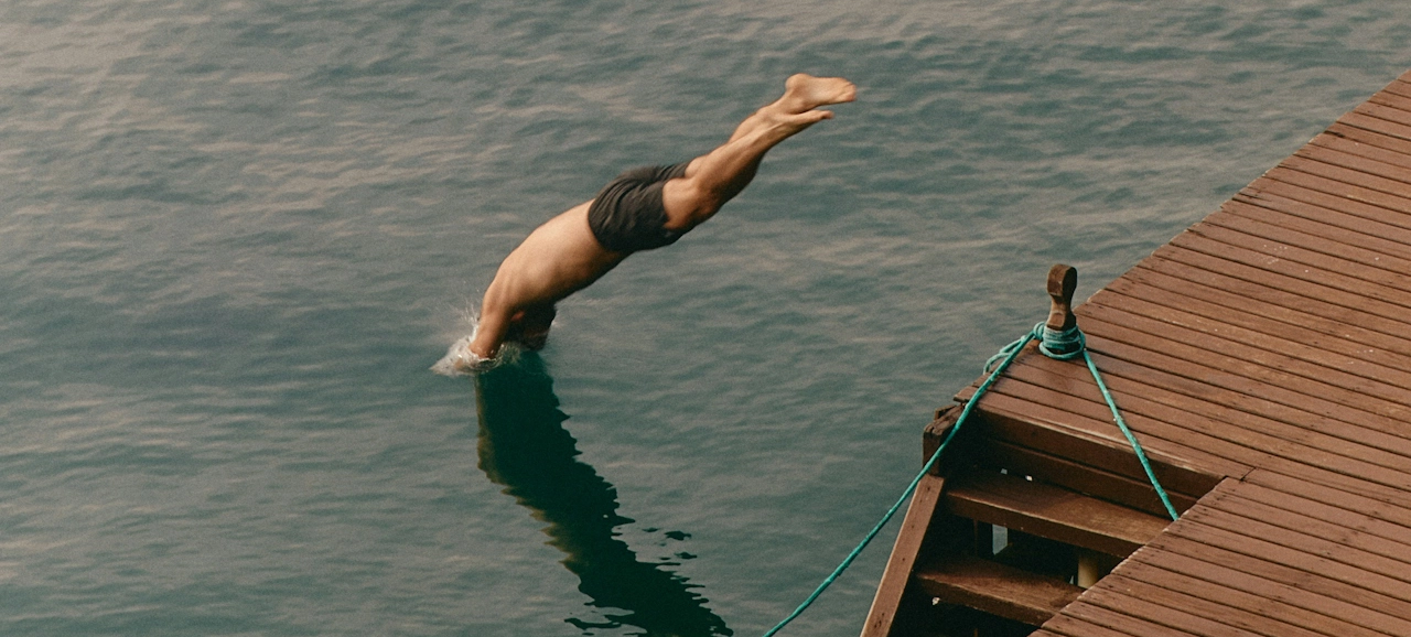 A person diving headfirst into a body of water from a wooden dock. The dock has ropes tied to posts, and the water appears calm.