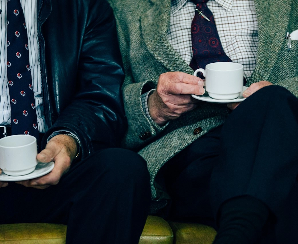 Two elderly men sitting side by side, each holding a white cup and saucer. The focus is on their hands and cups.