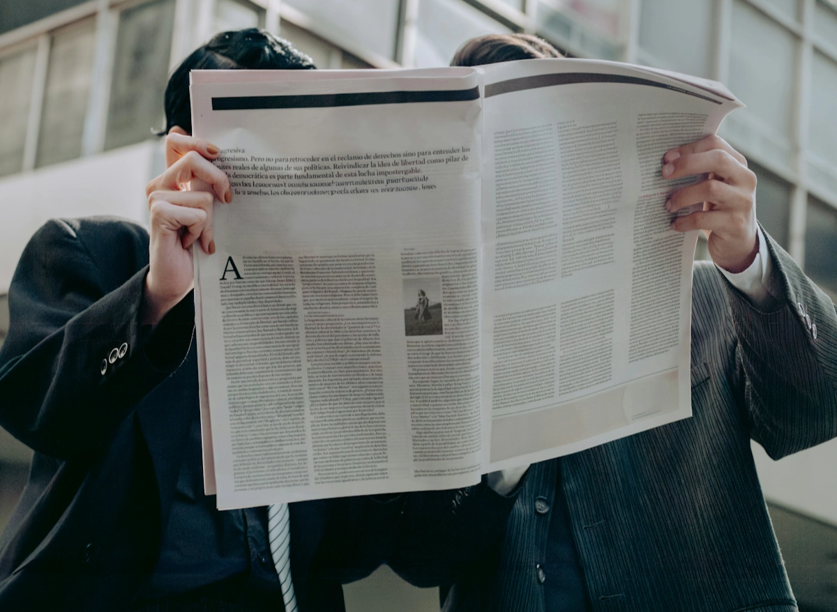Two business men in suits holding up a newspaper and reading it.