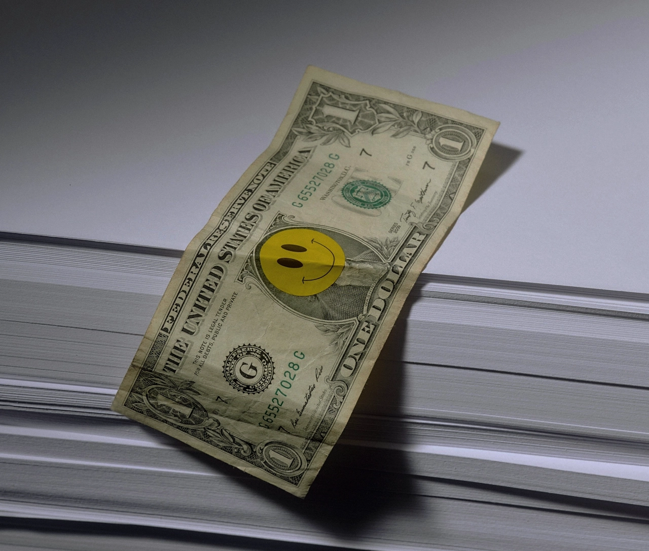 A dollar bill with a yellow smiley face sticker on it, placed on top of a large stack of white paper.